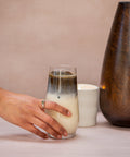 Hand holding a glass of layered drink next to a ceramic mug and wooden vase on a neutral background