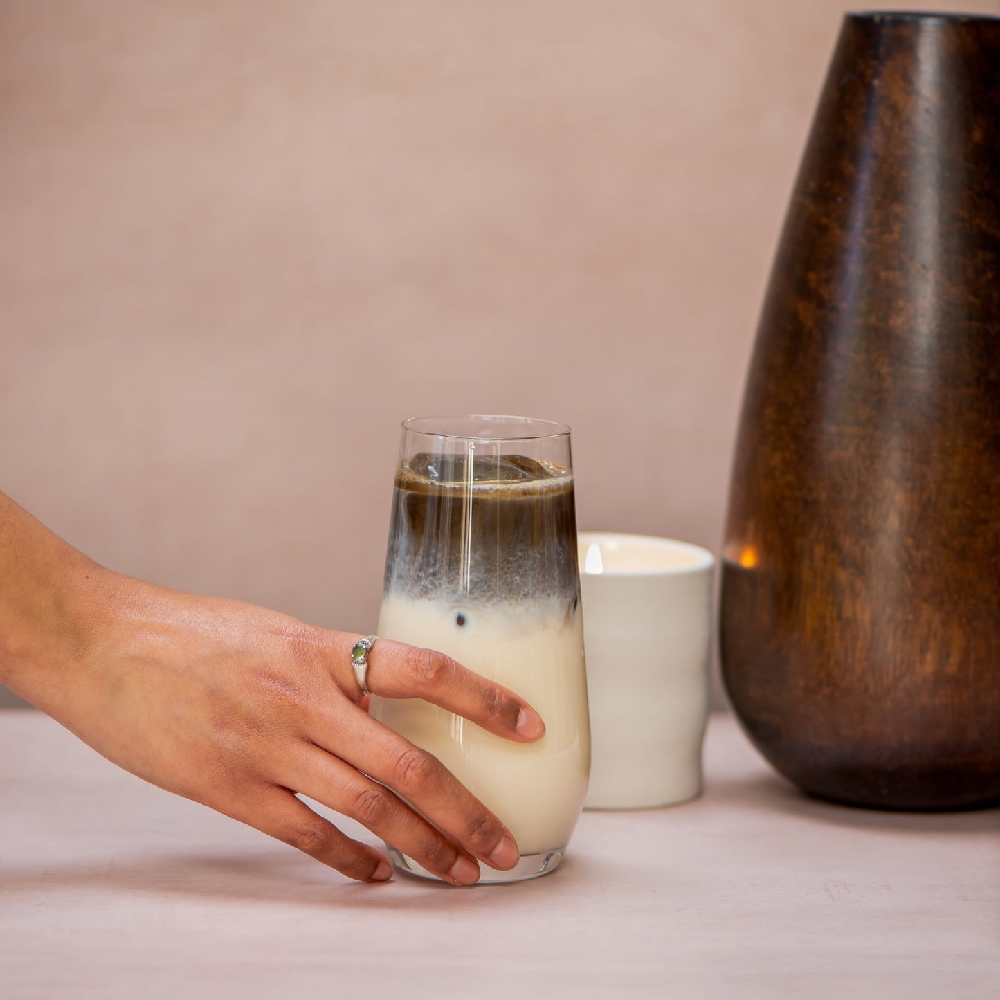 Hand holding a glass of layered drink next to a ceramic mug and wooden vase on a neutral background