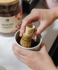 Blendsmiths bamboo whisk mixing House Choc into a smooth paste in a ceramic cup, with a House Choc storage jar behind