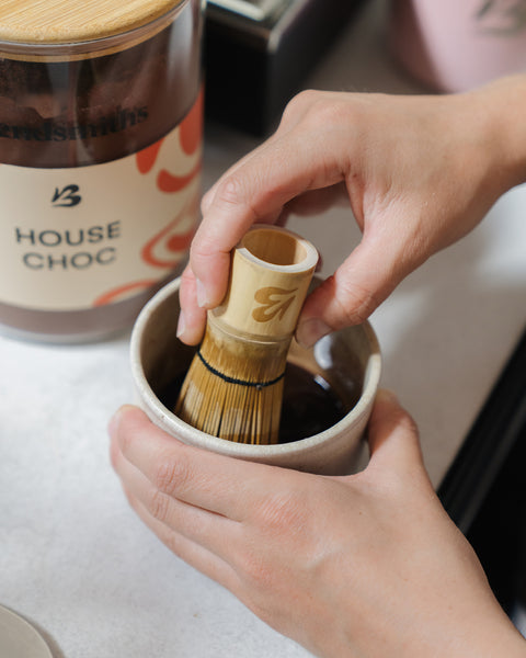 Blendsmiths bamboo whisk mixing House Choc into a smooth paste in a ceramic cup, with a House Choc storage jar behind