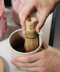 Close-up of a Blendsmiths bamboo whisk mixing a deep pink chai paste in a ceramic cup, with a Pink Chai storage jar and pink milk jug behind