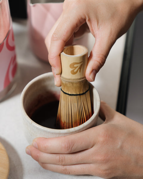 Close-up of a Blendsmiths bamboo whisk mixing a deep pink chai paste in a ceramic cup, with a Pink Chai storage jar and pink milk jug behind
